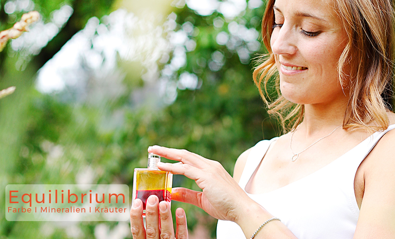 Aura-Soma Equilibrium Banner Eine glückliche Frau hält eine Aura-Soma-Flasche vor einem Baum in der Hand.